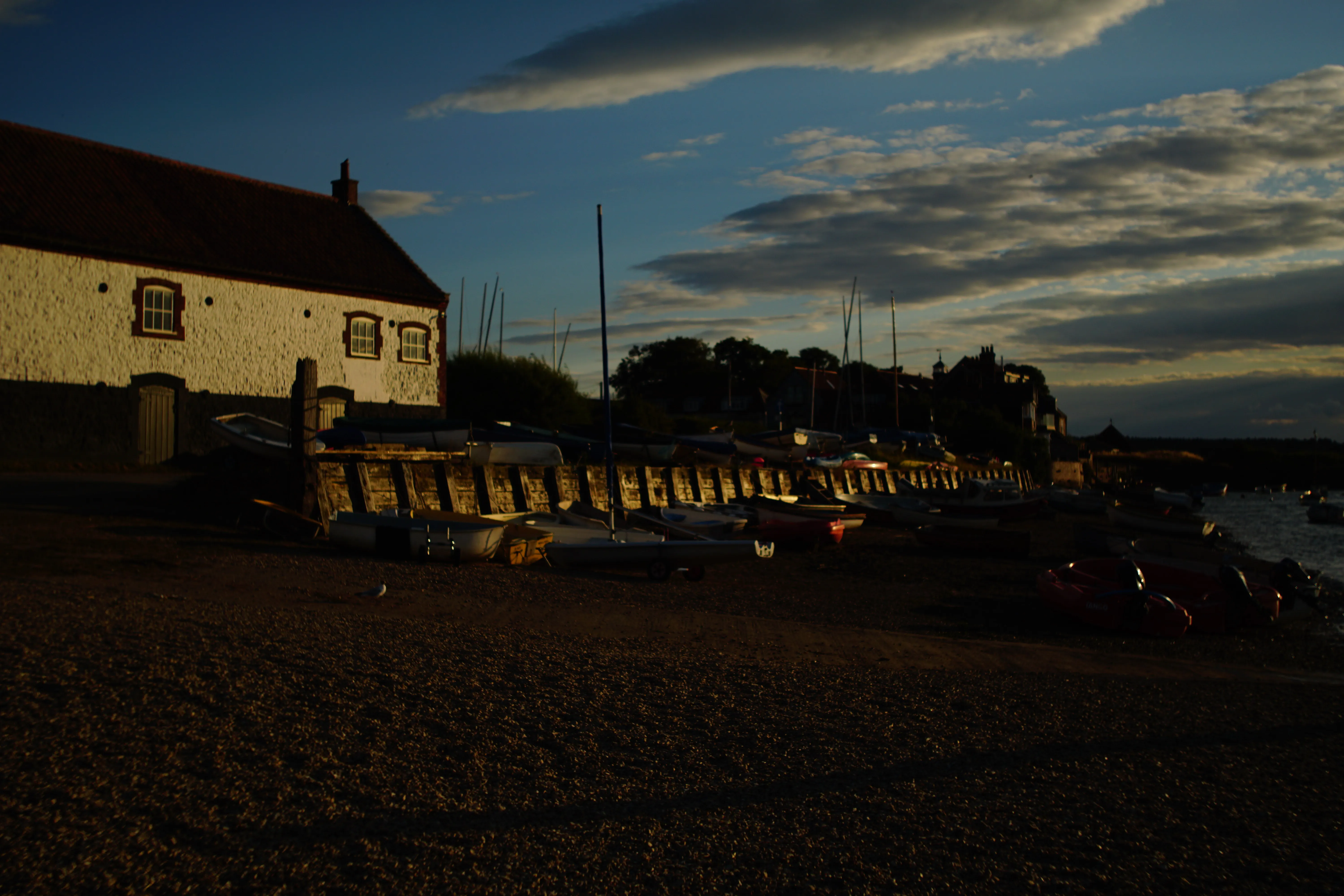 Low evening sun catching the flint boathouse at Burnham Overy Staithe, with dinghies drawn up on the shingle beside the creek.